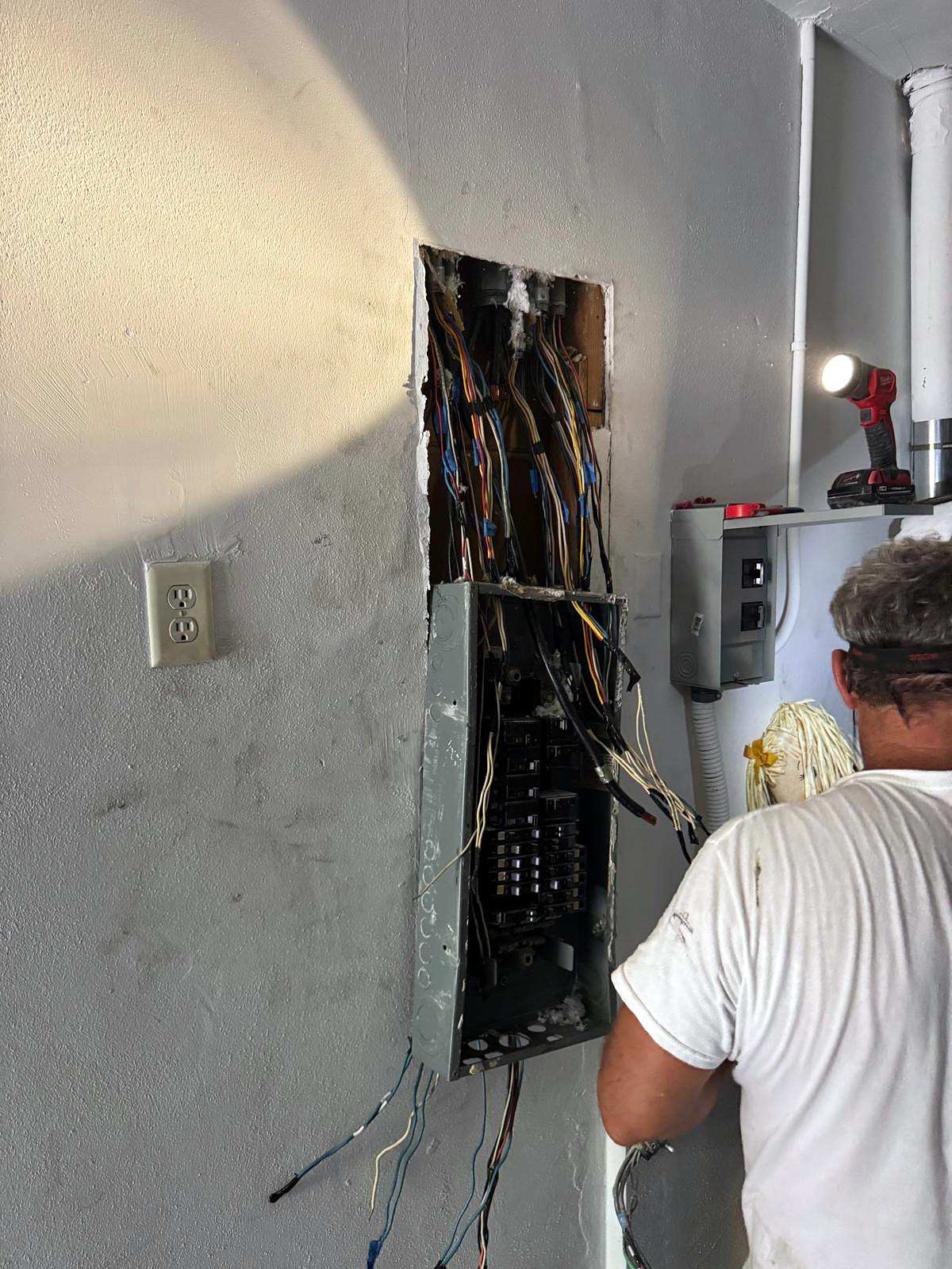 Electrician in a white T-shirt working at an open panel in a wall, wires fanned out around it