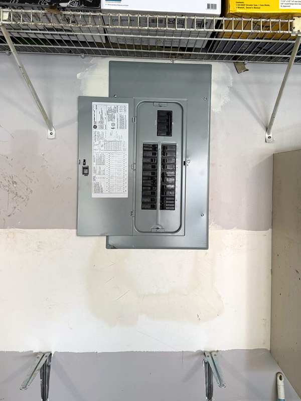 Older breaker panel with its door open on a garage wall, under a wire shelf